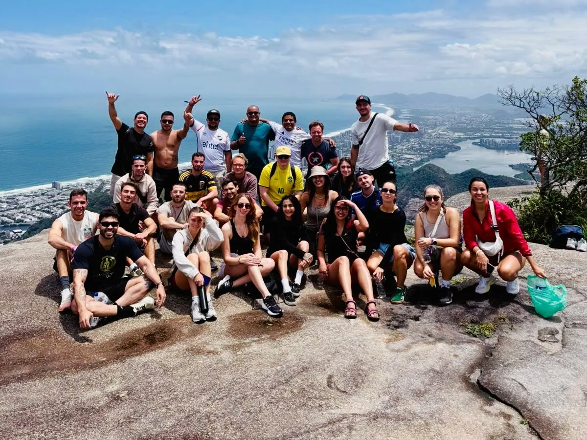 Alunos da Teacher Gabriela e viajantes de vários países no alto de uma trilha no Rio de Janeiro, com o mar ao fundo